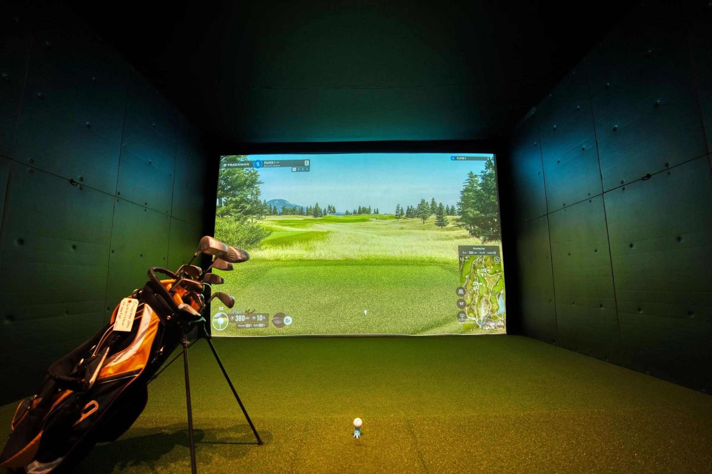Indoor golf simulator bay: Trackman screen showing a fairway, clubs bag in foreground, ball on tee
